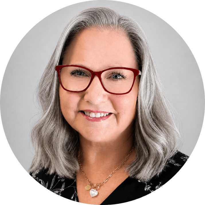 Catherine 1 - Visalia Recovery Center Portrait of a smiling woman with shoulder-length gray hair, red glasses, wearing a black patterned top and a gold heart pendant necklace.