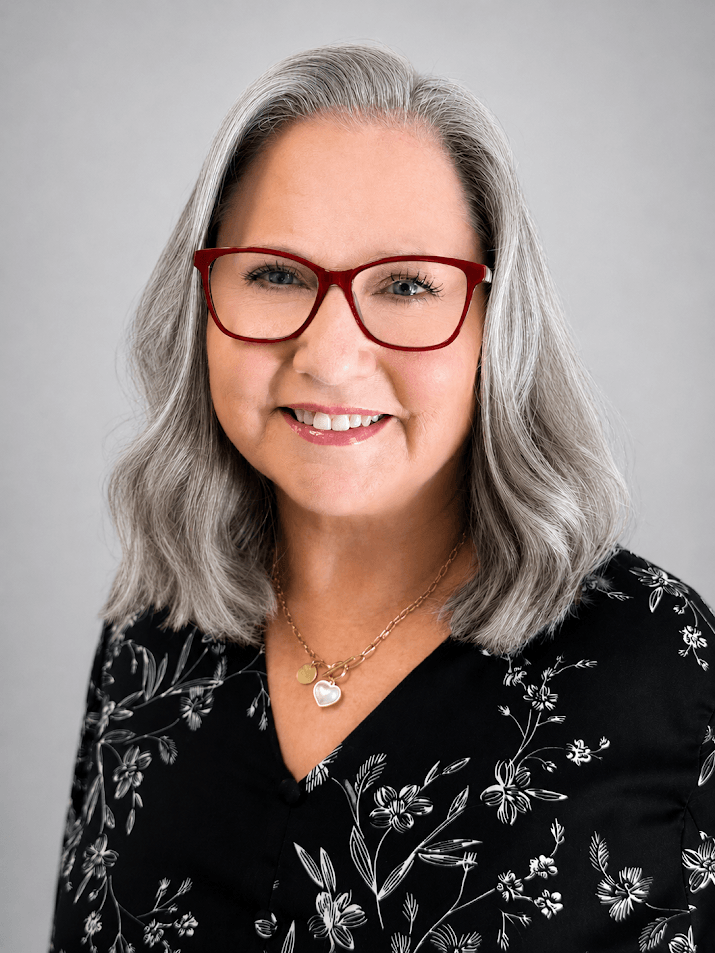 Smiling woman with shoulder-length gray hair and red glasses, wearing a black floral blouse and a heart pendant necklace.
