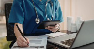 Healthcare professional in blue scrubs writes on a form with a pen, tablet in hand, beside a laptop and bottles on the desk.