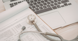 Stethoscope rests on a medical form on a clipboard beside a laptop keyboard in a bright workspace.