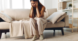 Woman sits on a beige sofa with hands on her head, appearing distressed in a bright living room setting.