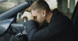 Man sitting in car with head bowed, resting on steering wheel, conveying stress or despair in a dim interior.