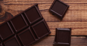 Dark chocolate bar with square sections on a wooden surface, with two broken pieces nearby on the right.