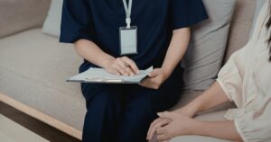 - Visalia Recovery Center Medical professional in navy scrubs with a clipboard and pen, seated on a couch with a patient for a consultation.