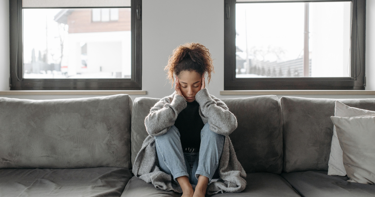 Young woman sits on a gray sofa with her hands on her temples, looking distressed in a living room with windows behind her.