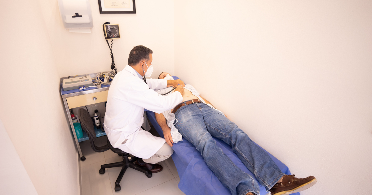 Doctor wearing a white coat and mask uses a stethoscope on a patient lying on an examination table in a small clinic room.